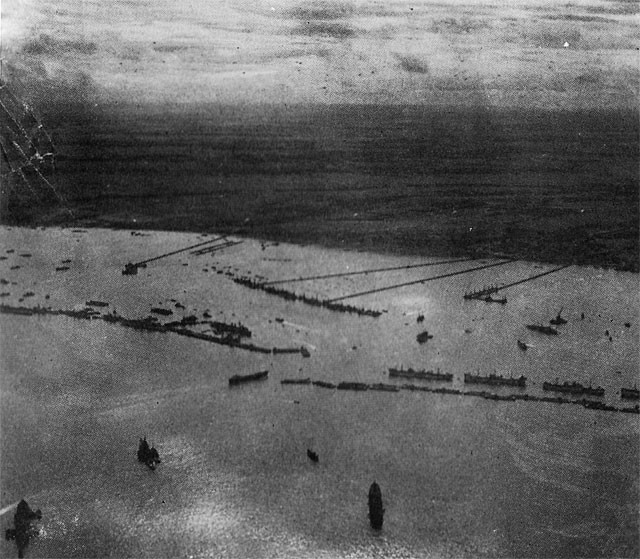 Here is a general view of the "MULBERRY" harbor completed and in full operation. In front is the breakwater of caissons and blockships; to the center is the row of pierheads forming the wharf, and running from them to the shore are the floating roadways. All together they form a port about as big as Dover.