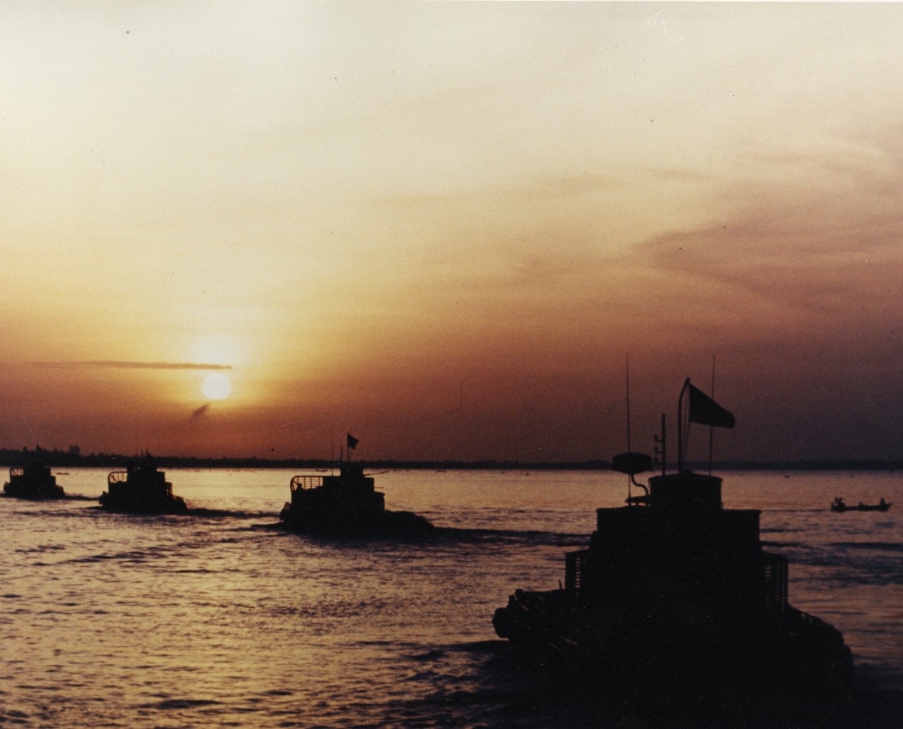 A line of boats moves along the water with a sunset behind them