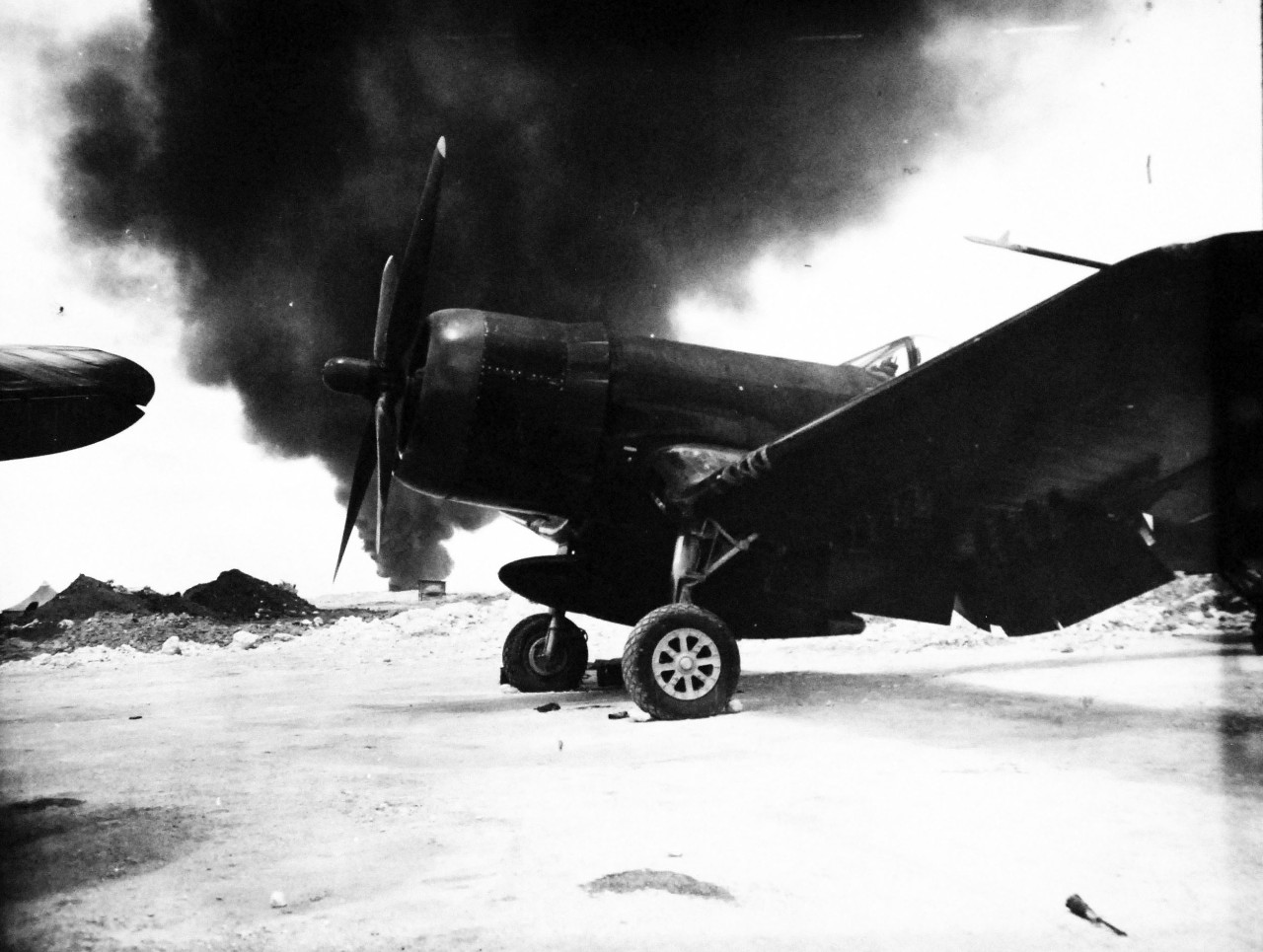 127-GW-524-125708:  Vought F4U Corsair, June 1945.  Against a background of heavy smoke which belched from a fuel dump fire on Okinawa, the F4U-4 “Corsair” fighter stands in its revetment.   Photographed by Corporal W.C. Beall, 19 June 1945. Official U.S. Marine Corps photograph, now in the collections of the National Archives. 