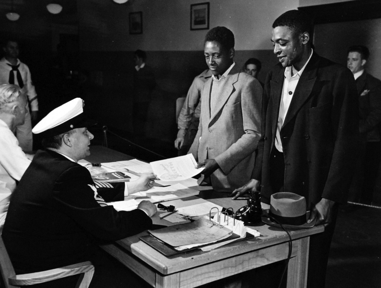 80-CF-882-2_Box 175: First African Americans Sworn in for Clerical Service, 1942.   Original caption, “First African-Americans Sworn in for Clerical Service.”  Walter Hurtt, accompanied by his father Wyoming Hurtt, gets enlistment data at U.S. Navy Recruiting Station, 67 Broad Street, New York.  Hurtt was one of the first African-Americans to apply under the Navy’s new rule allowing African-Americans to enlist in other than mess attendant’s capacity.  Possibly summer of 1942.  Official U.S. Navy photograph, now in the collections of the National Archives Note, this photograph has not been fully verified.   