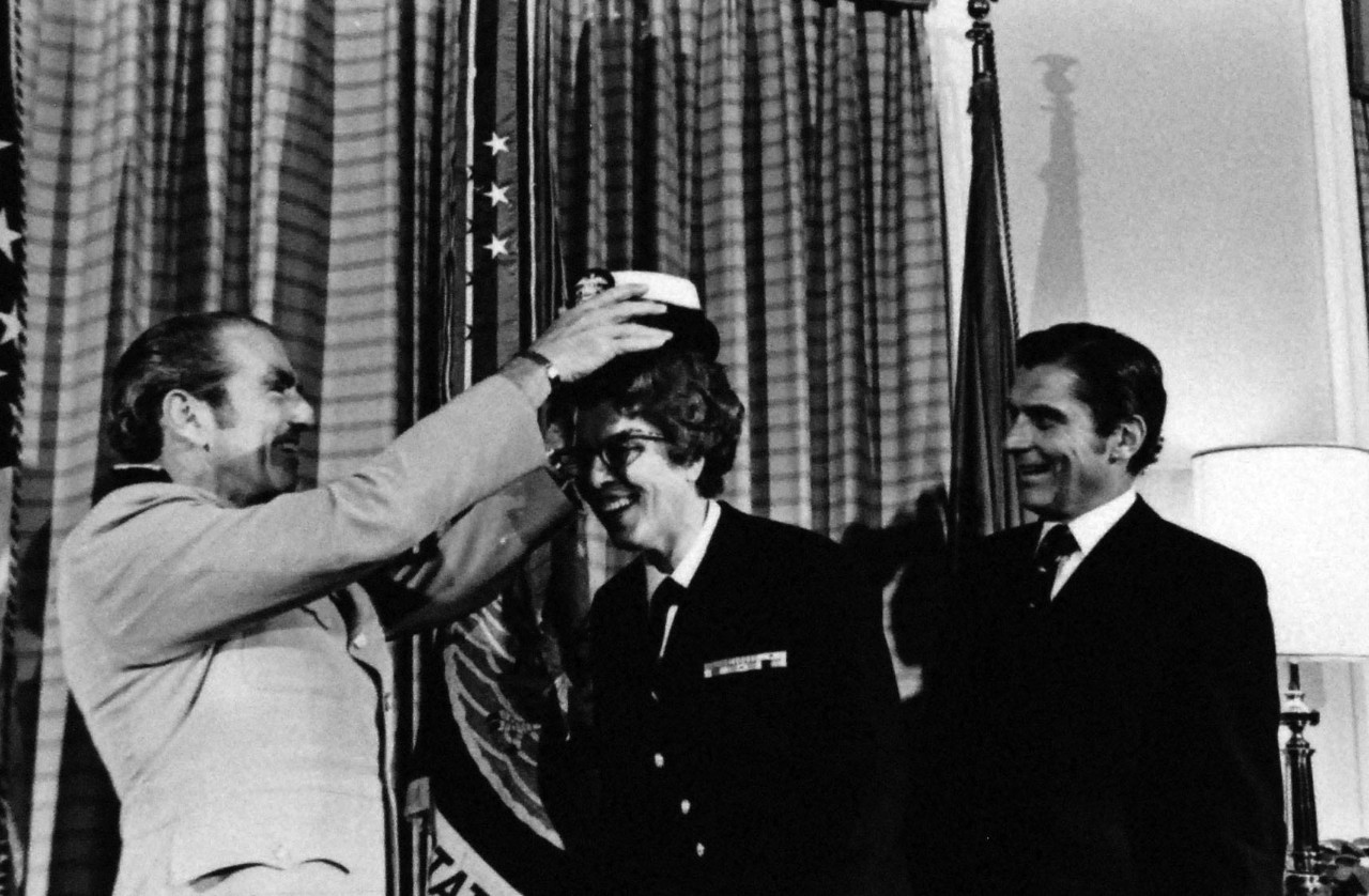 428-GX-USN 1151695:   Rear Admiral Arlene B. Duerk, June 1972.   Washington, D.C.  Chief of Naval Operations Admiral Elmo R. Zumwalt puts on her new Admiral’s combination hat on the head of  Rear Admiral Arlene B. Duerk on her promotion during a ceremony in the Office of the Secretary of the Navy John W. Warner who looks on t right.  Rear Admiral Duerk, Director of the Navy Nurse Corps was the first woman to reach the rank of Rear Admiral.   Photographed by PH1 Billy L. Mason, June 1, 1972.    Official U.S. Navy Photograph, now in the collections of the National Archives. Photographed from small reference card.   