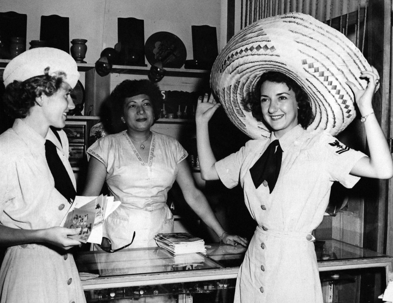 USN 709023:    Selecting interesting souvenirs of moderate price is one of the privileges of world travelers, October 1953.  HM2 Eileen Paluzzi tries on a hand-made beach hat, which she decided not to buy.   HM3 Maria Myers bought some postcards to send to the folks back home.    Photograph released October 26, 1953.   Master Caption:   Navy Accepts Women Volunteers for Duty on the High Seas.   For the first time in history, the U.S. Navy has accepted volunteers for duty on the high seas from women in the enlisted WAVES.  Those eligible were WAVES of the Hospital Corps to fill sixty-three billets on ships of the Military Sea Transportation Service.   Official U.S. Navy photograph, now in the collections of the National Archives.   