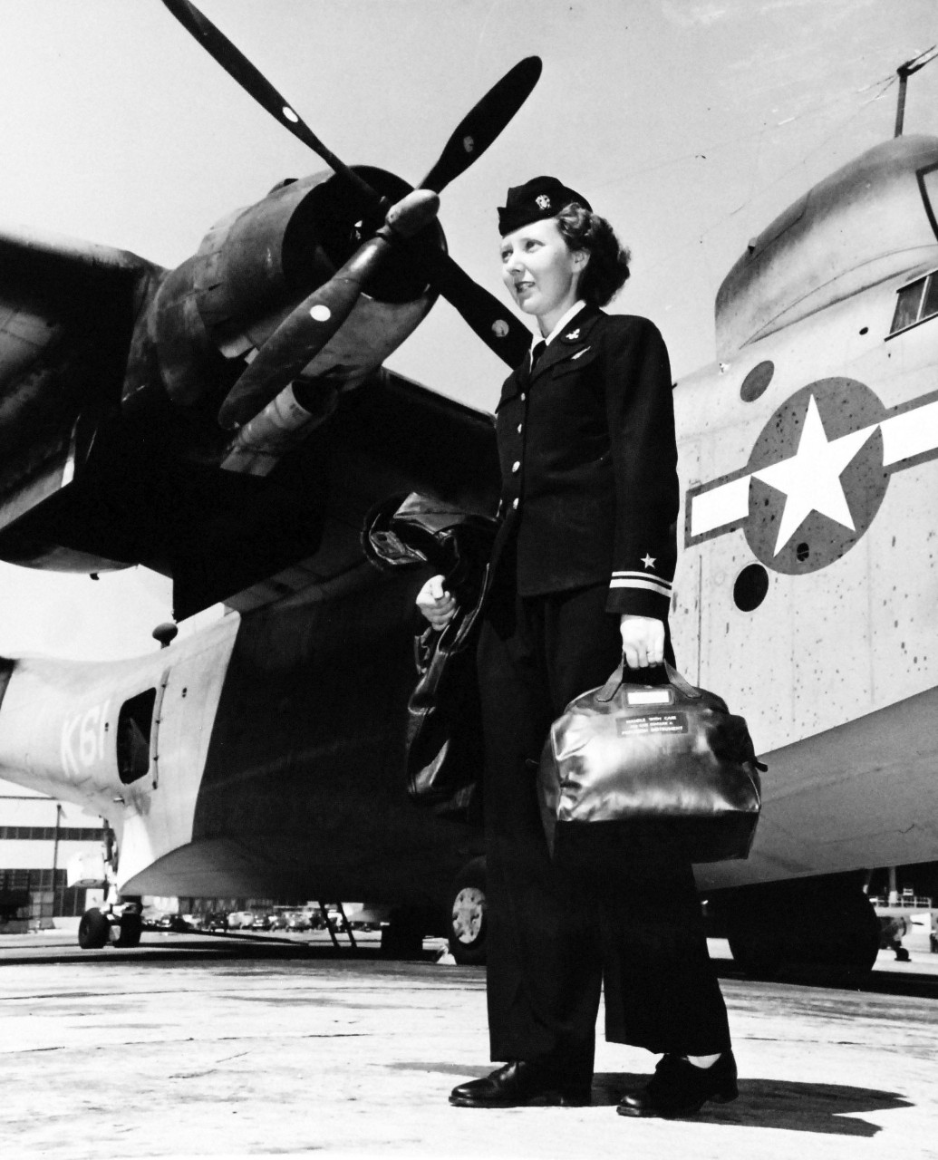 80-G-49793: Service Dress Blue, April 1945.   Lieutenant Junior Grade Kathleen K. Ballard, Naval air navigator, walks to her plane at the Naval Air Station, Alameda, California.  With her, she carriers her flight clothing and in the bag is a sextant which she will use in flight to obtain “fixes”.  Plane in the background is a PBM.   Received April 7, 1945.   Official U.S. Navy photograph, now in the collections of the National Archives.