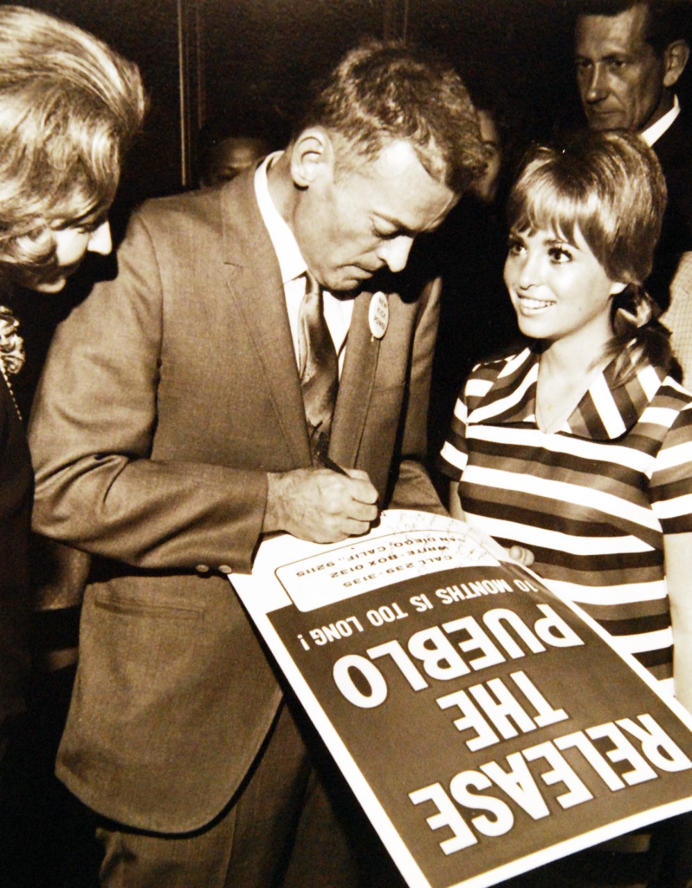 428-GX-USN-1140174:   San Diego, California, January 12, 1969.   Commander Lloyd M. Bucher, Commanding Officer of USS Pueblo (AGER-2), signs an autograph for a Hollywood starlet on a “Release the Pueblo” poster during a part at the La Baron Hotel.  Mrs. Bucher is at left.  Photographed by PH1 Robert E. Woods.   Official U.S. Navy Photograph, now in the collections of the National Archives.  (2017/05/23).  Photographed from reference card.  