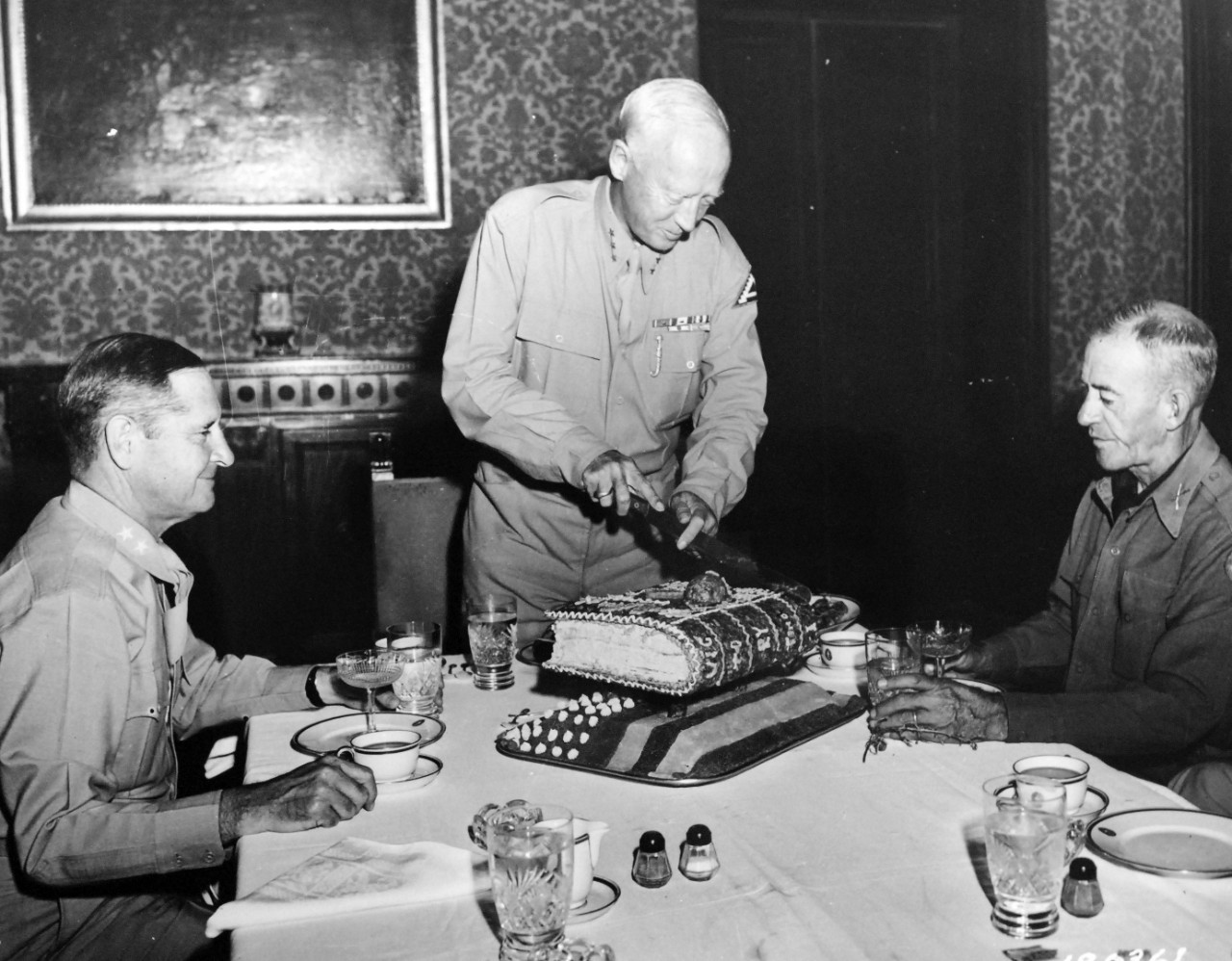 111-SC-180761:  Operation Husky, July-August 1943.   Lieutenant General George S. Patton, Jr. cuts victory cake baked on the occasion of the victory of General Patton’s Seventh Army in Sicily.  Left is Major General Geoffrey Keys, Deputy Commander of the Seventh Army and Colonel Paddy Flint, Commanding Officer of a regiment in the Ninth Infantry Division, is seated at right, Sicily, September 1943.  U.S. Army Signal Corps Photograph.   Courtesy of the Library of Congress: Lot-6040-9. (2017/04/21).  