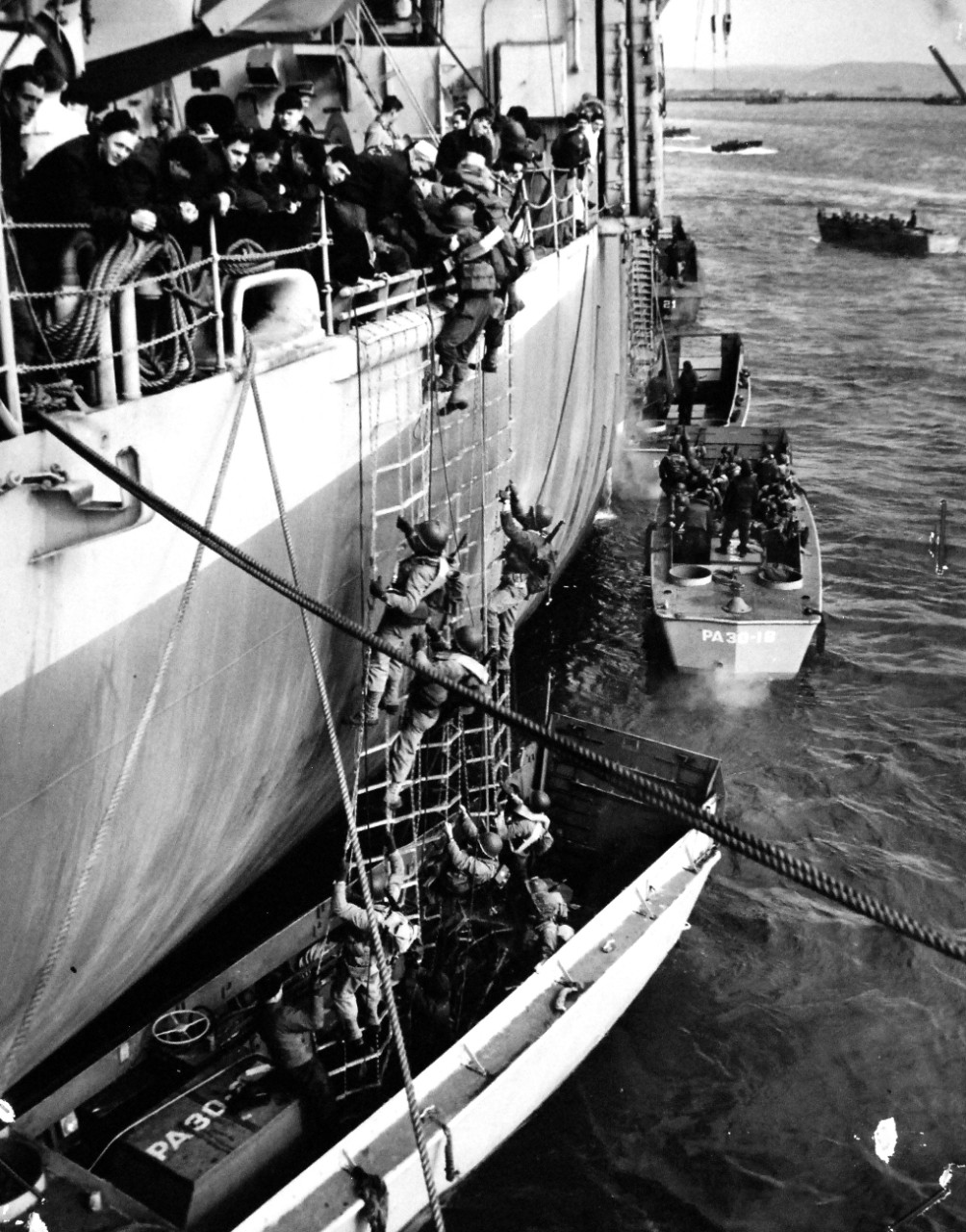 26-G-3433:  Normandy Invasion, June 1944.  Up the Landing Nets.   USS Thomas Jefferson (APA-30), at a British port and “sea taxis”, landing craft, swarm alongside packed with troops.  Up the landing nets go its fighters and over the side with Coast Guardsmen giving the boys a hand, June 1944.   Official U.S. Coast Guard Photograph, now in the collections of the National Archives.   (2015/5/19).  