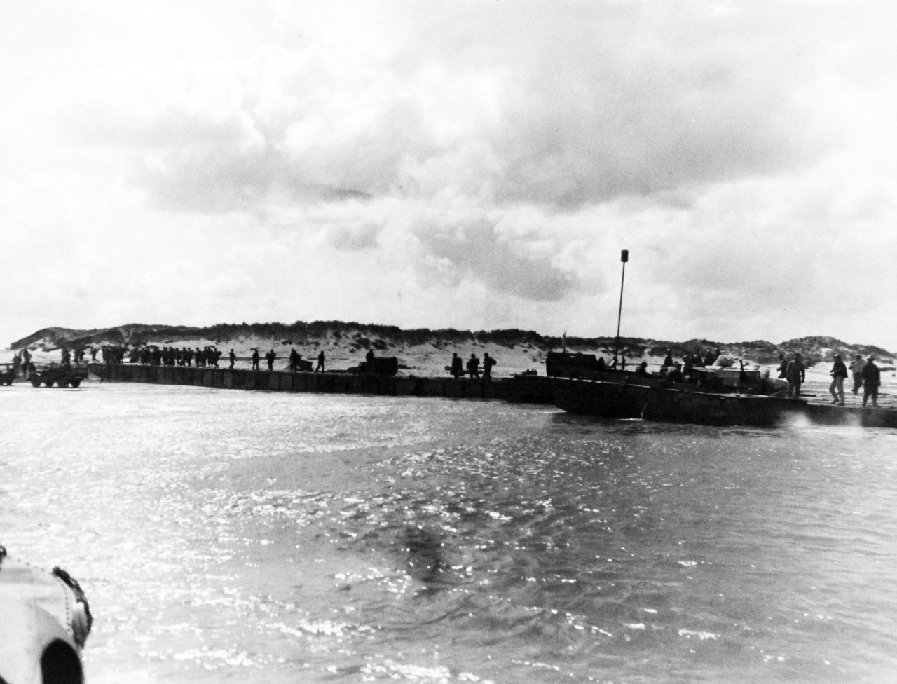 80-G-285126:  Normandy Invasion, Mulberries, June 1944.   The pontoon causeway at “Utah” beach, forming part of the U.S. Mulberry the man-made harbor off coast of France, built to protect landing craft from storms in English Channel.   Shown:  Reinforcing troops march over causeway.  Low tide leaves some sections of the latter dry.  Photograph released November 7, 1944.  U.S. Navy photograph, now in the collections of the National Archives.  (2016/03/15).