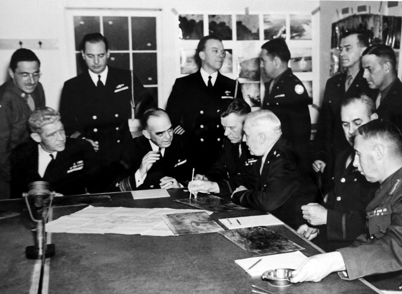 LC-Lot-803-3:  Aleutian Islands Campaign, June 1942 - August 1943.   Allied Invasion of Kiska, August 15-24, August 1943.   American and Canadian military leaders in the North Pacific are shown here holding a final conference at the Aleutian headquarters of Vice Admiral Thomas C. Kinkaid, Commander, North Pacific Force, before the Allied landing on Kiska, August 15, 1943.   Seated around the table in Admiral Kinkaid’s headquarters are, (left to right):  Rear Admiral Francis W. Rockwell, USN, Commander, Pacific Amphibious Forces; Vice Admiral Thomas C. Kinkaid, USN, Commander, North Pacific; Major General G.H. Corlett, USA; Lieutenant General Simon B. Buckner, USA; Commanding General, Eleventh Air Force; Major General G.R. Parkes, RCA, General Officer, Commanding-in-Chief, Canadian Pacific Command.   Standing, (left to right): An unidentified Army officer; Commander R.K. Dennison, USN, Chief of Staff to Rear Admiral Rockwell; Captain O.S. Colclough, USN, Chief of Staff to Vice Admiral Kinkaid ; Colonel Carl T. Jones, AUS, Chief of Staff to Major General Corlett; Brigadier General J.L. Ready, USA, and Brigadier General E.D. Post, AUS, Chief of Staff to General Buckner.   U.S. Navy photograph.  Photographed through Mylar sleeve.  Courtesy of the Library of Congress.  (2015/11/06).