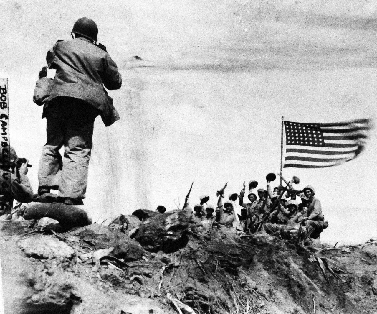 127-GW-319-112720: Iwo Jima Operations, February 1945.   Victors – In the stiff breeze atop Mount Suribachi, Iwo Jima, “Old Glory” whips against the sky as cheering Marines raise their voices and weapons in the historic moment for posterity.  Note, Joe Rosenthal, photographer is in the left foreground.   Photographed by Bob Campbell, February 23, 1945.   Official U.S. Marine Corps Photograph, now in the collections of the National Archives.  (2016/09/06).