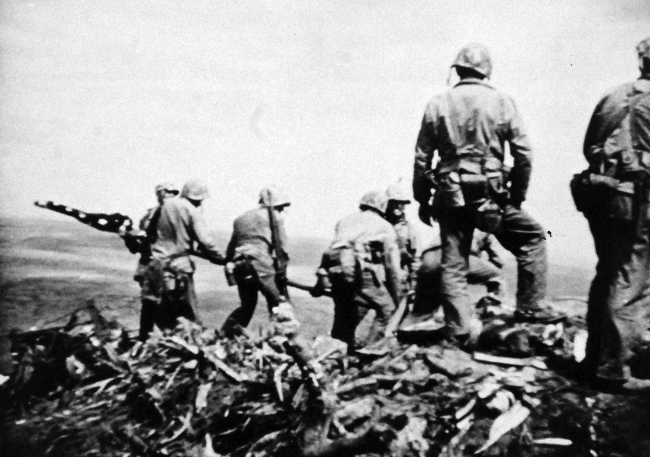 127-GW-319-116770:   Iwo Jima Operations, February 1945.  Over Suribachi.  A “still” taken from the 16mm movie series of the U.S. Marines raising the American flag on the summit of Mount Suribachi, Iwo Jima.     Photographed by Genaust, February 23, 1945.   Official U.S. Marine Corps Photograph, now in the collections of the National Archives.  (2016/09/06).