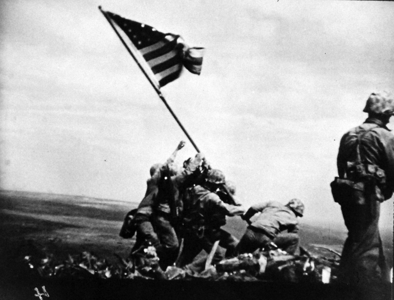 127-GW-319-116774: Iwo Jima Operations,  February 1945.  Over Suribachi.  A “still” taken from the 16mm movie series of the U.S. Marines raising the American flag on the summit of Mount Suribachi, Iwo Jima.     Photographed by Genaust, February 23, 1945.   Official U.S. Marine Corps Photograph, now in the collections of the National Archives.  (2016/09/06).