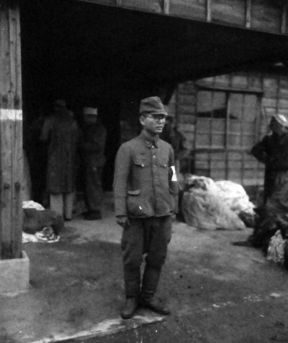 80-G-339282:  Prisoner of War Camp, Omori Prison Camp, Toyko, Japan, 1945.    Shown here is Japanese official with liberated prisoners in the background, September 1, 1945.     Omori was one of Tokyo, Japan, biggest wards. Image taken by Ensign Simon and Photographer’s Mate Second Class Colb.  Official U.S. Navy photograph, now in the collection of the National Archives. (2013/08/14).    Note, the original photograph is about 1” x 1” in size.   
