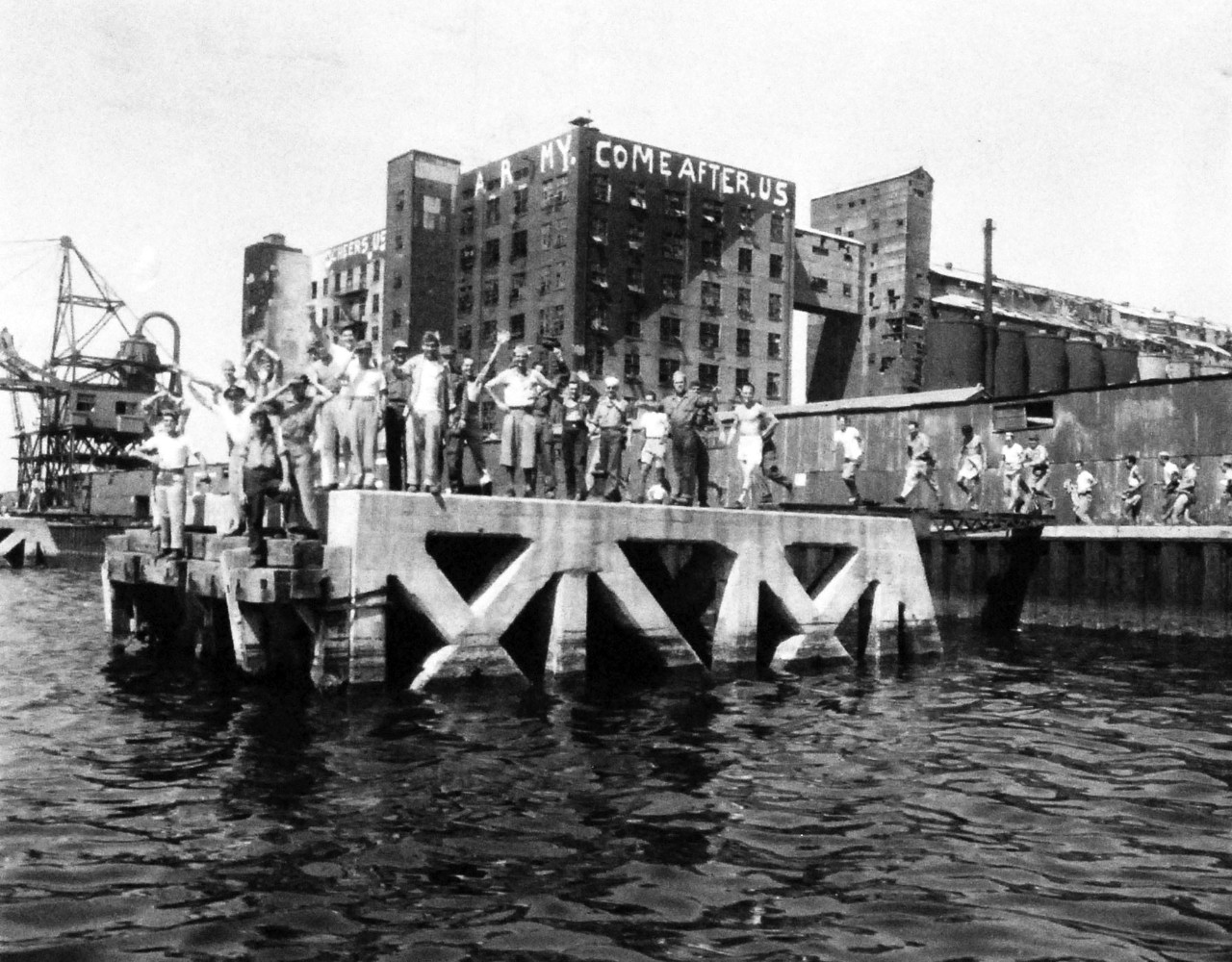 80-G-490450:    Allied Prisoners of War, Tokyo, Japan.   Prisoners of war wait on a small dock in the Tokyo waterfront area for rescue craft manned by the U.S. Navy to approach and taken them out to a waiting hospital ship, USS Benevolence (AH-13).   Tall structures in the background have been bedecked with signs calling for rescue, Sepember 1, 1945.  Official U.S. Navy photograph, now in the collections of the National Archives.   (2014/5/29). 