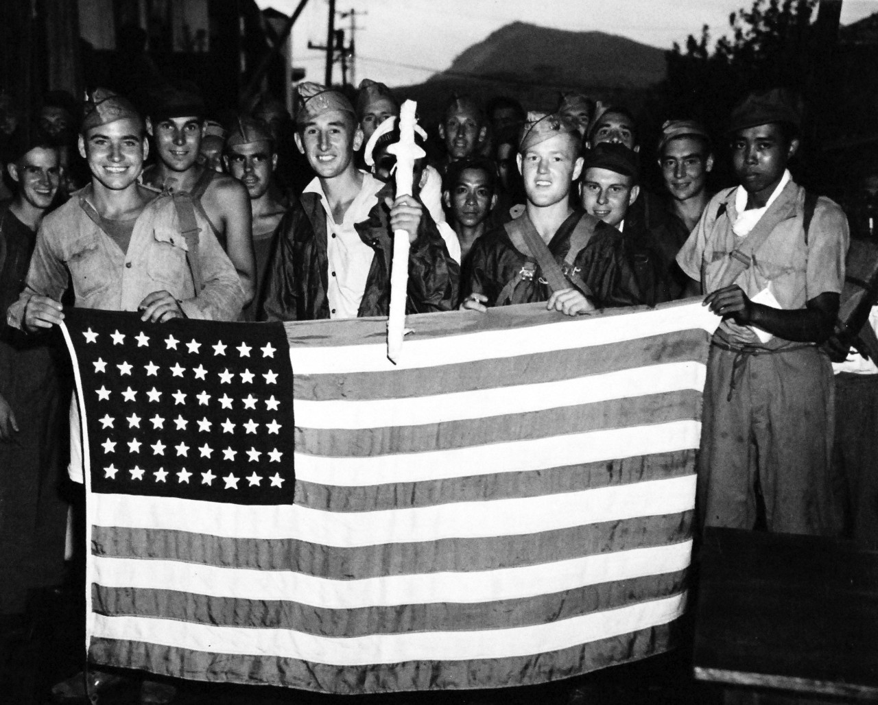 80-G-495671:   Allied Prisoners of War, Japan, 1945.   This United States Flag, held by grinning Allied Prisoners of War of the Japanese, was made from scraps of parachutes dropped by U.S. Navy and Army planes in mercy flights over the camps in the Nagasaki area of Japan.   The fliers dropped food, clothing and medical supplies to the prisoners who were rescued 13 September by mercy squads of the U.S. Navy.   Photograph released October 1, 1945.   Official U.S. Navy photograph, now in the collections of the National Archives.  (2014/05/29).    The original is a small photograph.   