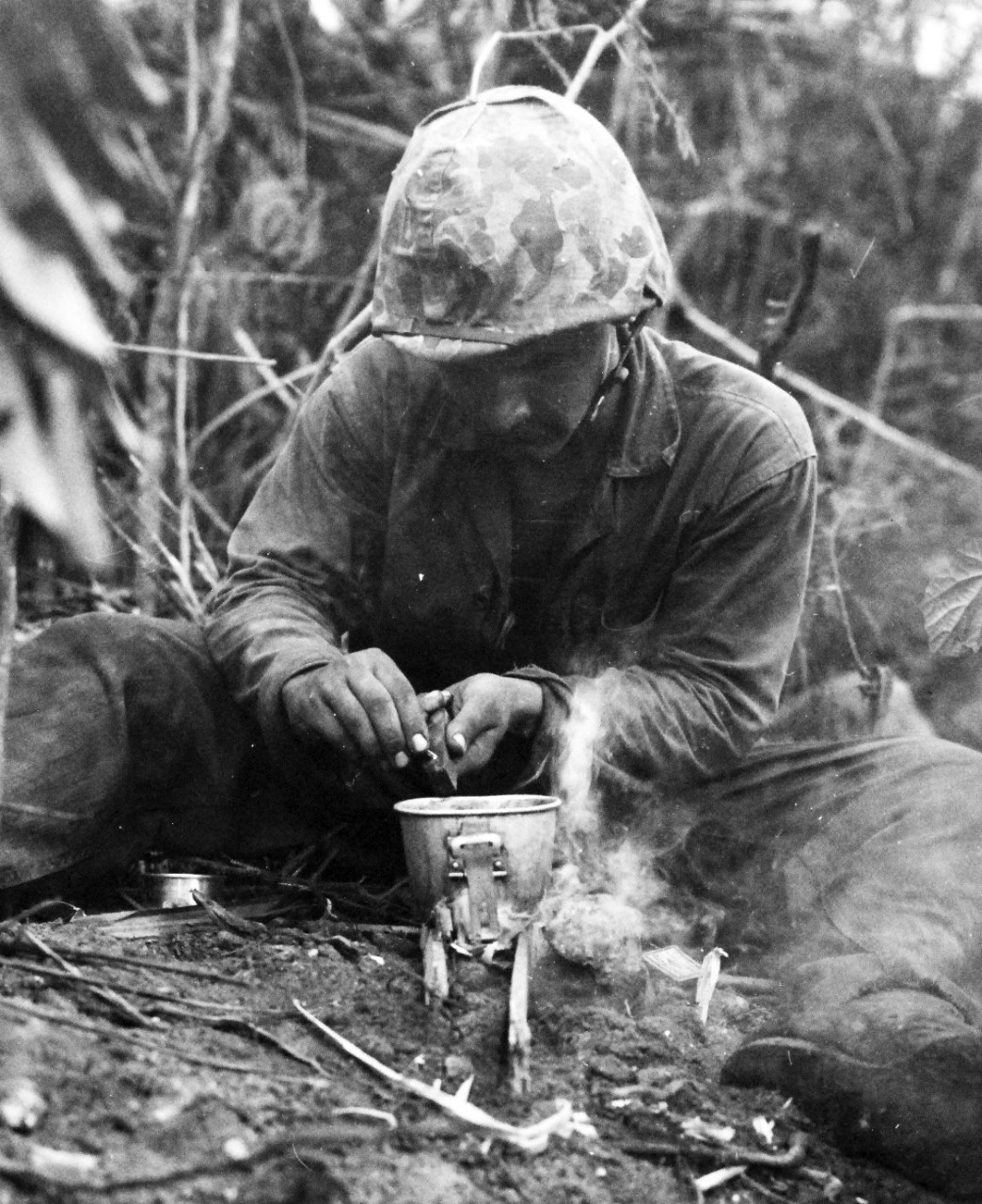 80-G-239318:   Invasion of Guam, July 21-August 10, 1944.   Private First Class Attilio Lattanz, who landed on Guam with the U.S. Marines on D-Day, has his first hot chow, several days later, as he scrapes chocolate from a ration bar into his mess cup, which he places on a small fire.  It wasn’t eight course dinner, but it was better than cold rations,   released August 5, 1944.    Official U.S. Navy Photograph, now in the collections of the National Archives.  (2017/03/07).