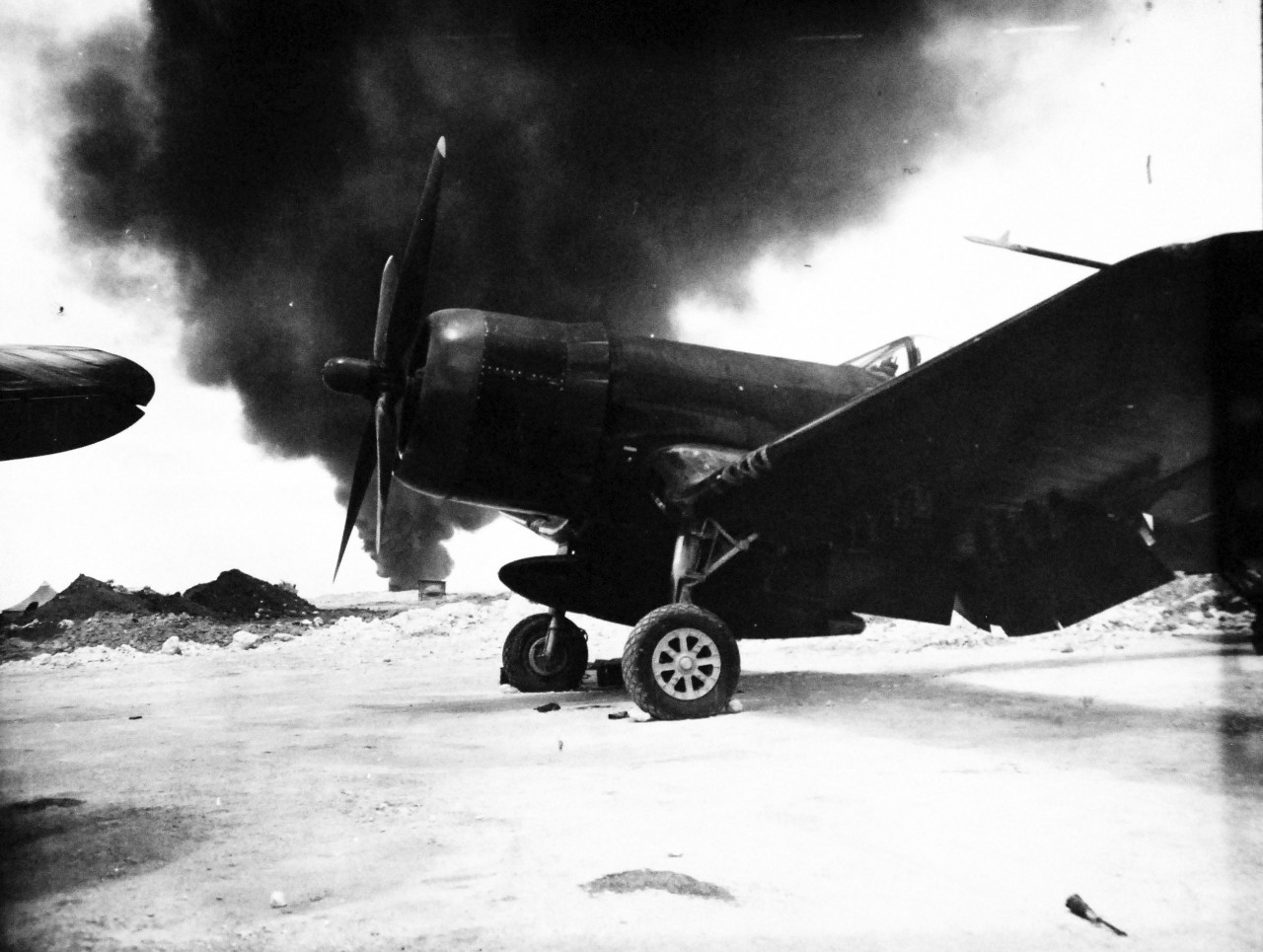 127-GW-524-125708:  Okinawa Campaign, April-June 1945.   Against a background of heavy smoke which belched from a fuel dump fire on Okinawa, the new F4U-4 “Corsair” fighter stands in its revetment.   Photographed by Corporal W.C. Beall, 19 June 1945.  Official U.S. Navy Photograph, now in the collections of the National Archives.   (2014/6/25). 