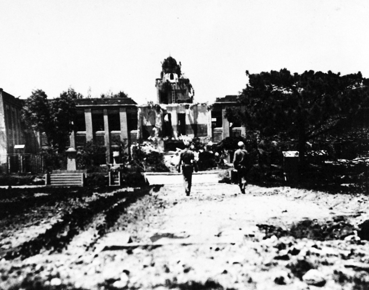 80-G-322706: Okinawa Campaign, April-June 1945.   Two First Division Marines walk along a rubble strewn road towards the ruins of the Japanese Naval Academy on Okinawa, Ryukyus Islands. The building was wrecked during pre-invasion bombardment, released April 22, 1945.  Official U.S. Navy photograph, now in the collections of the National Archives.  (2016/04/26).  