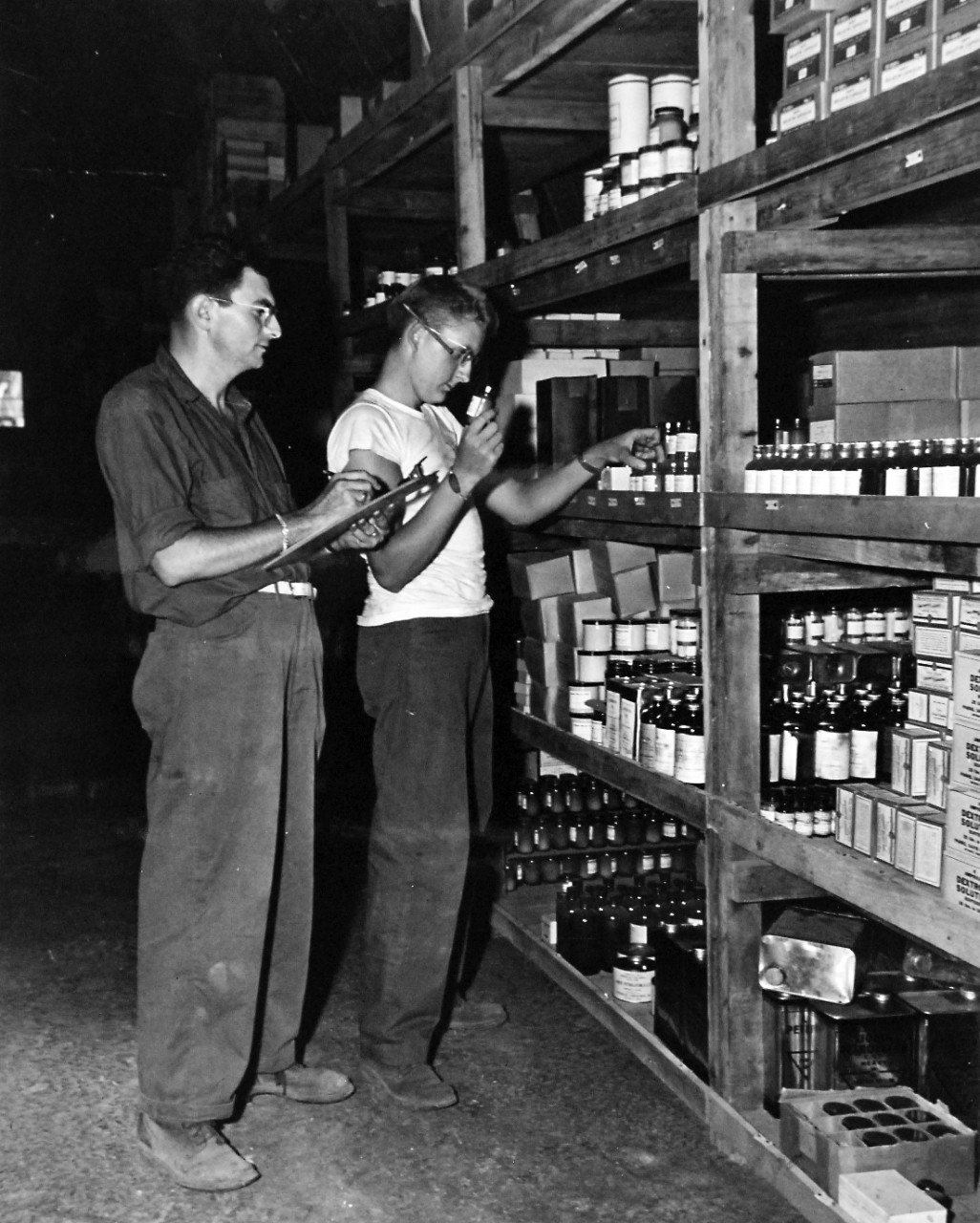 80-G-49580: Okinawa Campaign, April-June 1945.  Progress of construction on Okinawa. Left to right:  PhM1/C Albert Smolens and HA1/C Robert E. Murray taking inventory of supplies (from pins to wheelchairs) in a warehouse with 8000 square feet of shelves made from salvaged lumbar.   Photograph received June 29, 1945.   Official U.S. Navy photograph, now in the collections of the National Archives.  (2017/01/10).