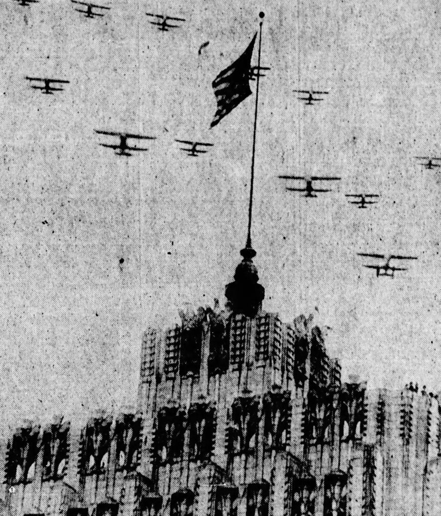 A group of bi-planes flying over a building with an American flag displayed from the spire. 