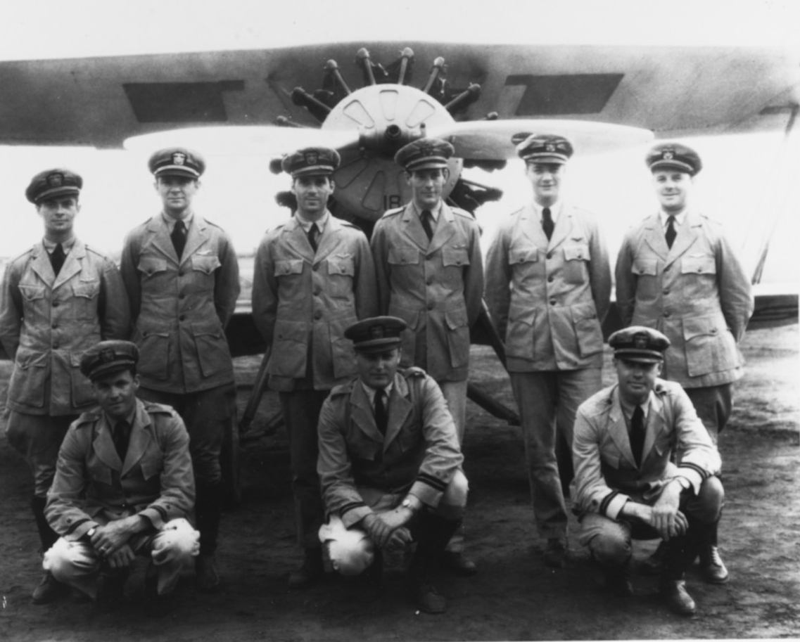Nine pilots in uniform pose in front of a parked biplane.