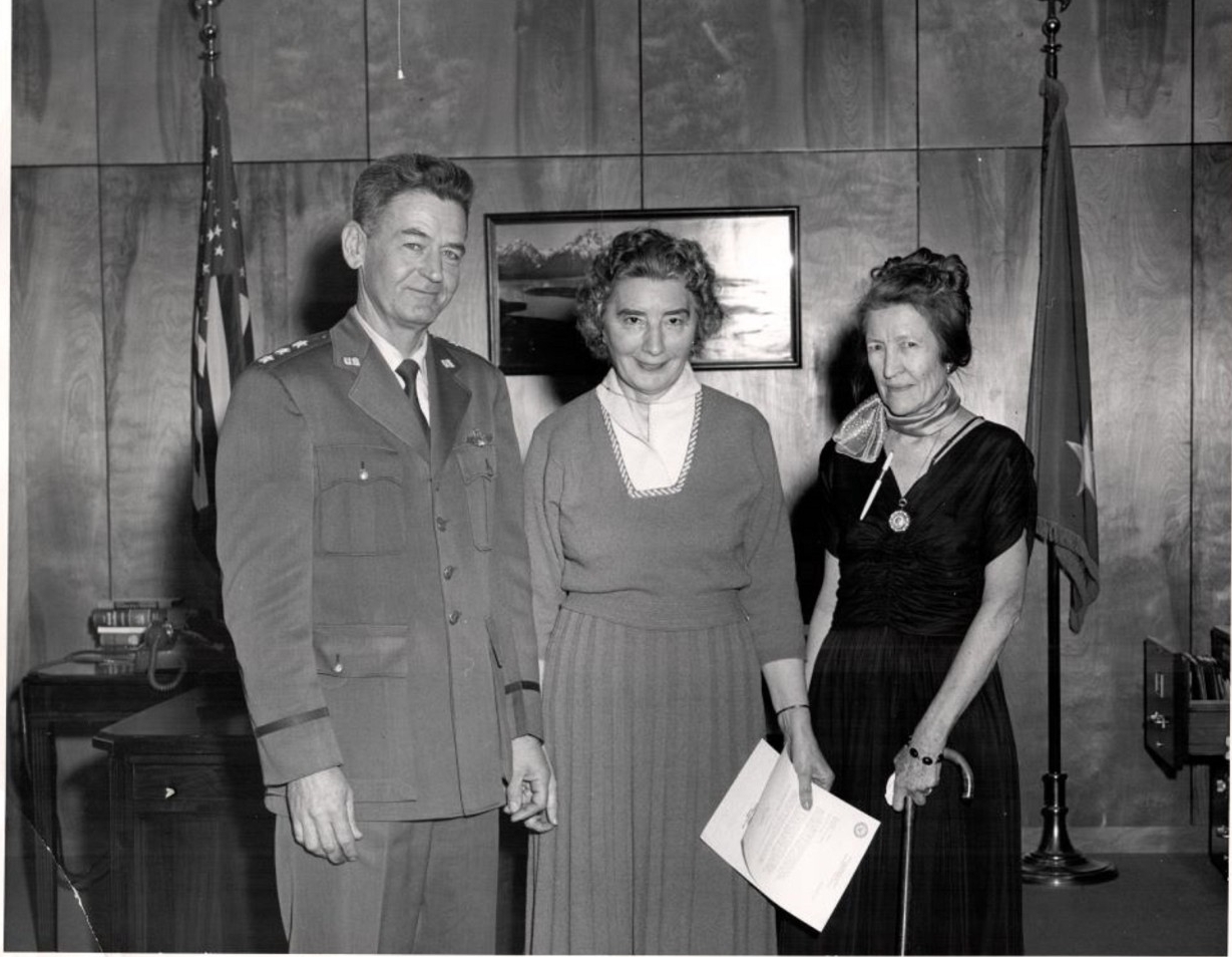 A man in uniform stands next to two women in dresses.
