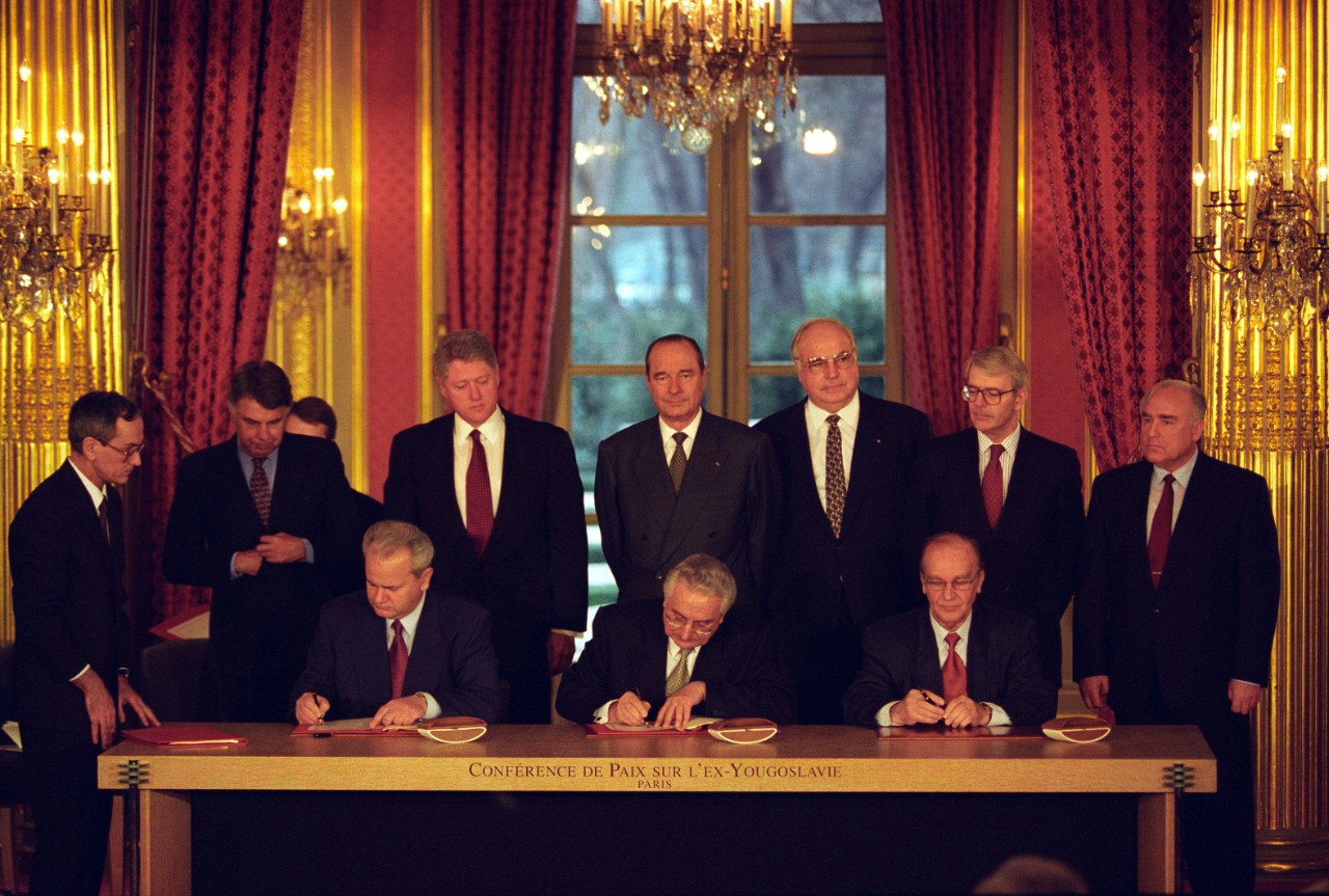 Three men seated at a table signing the document surrounded by eight other men. 