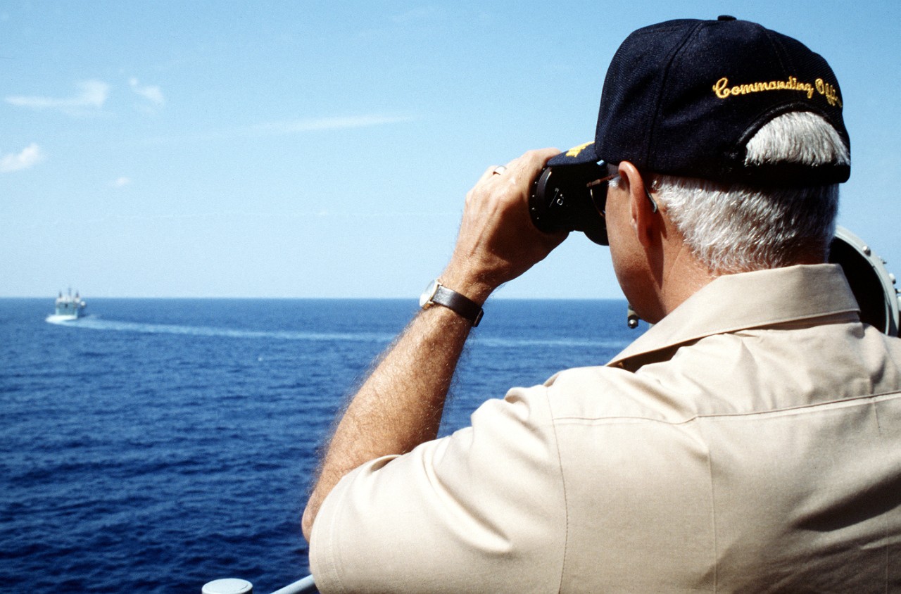 Man looks through binoculars to watch a small ship in the distance. 