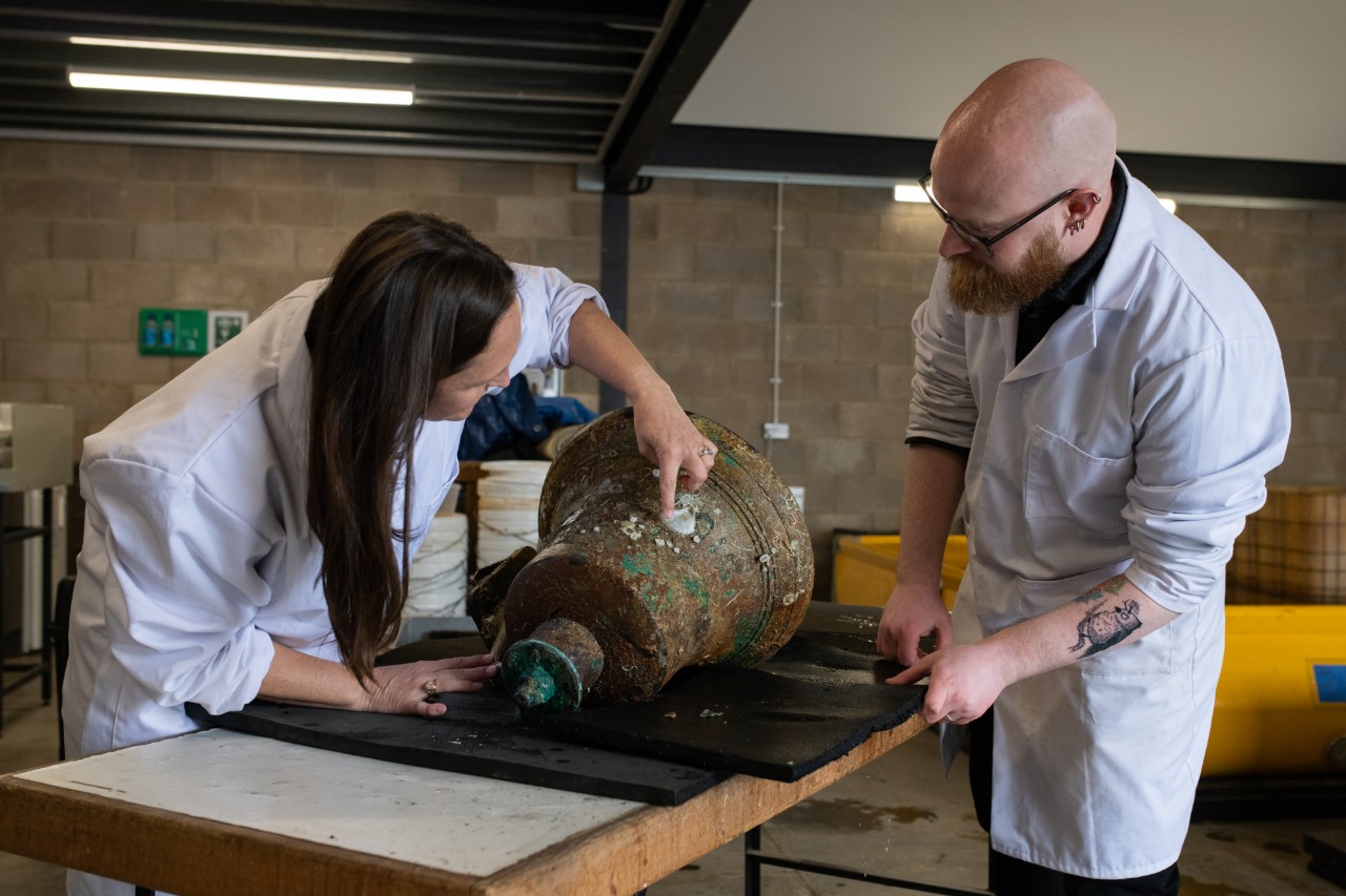 Shanna Daniel, lead archaeological conservator at Naval History and Heritage Command and Thomas Wicks, conservator at Wessex Archaeology, prepare the ship’s bell of USS Jacob Jones (DD 61) for conservation at Wessex Archaeology.