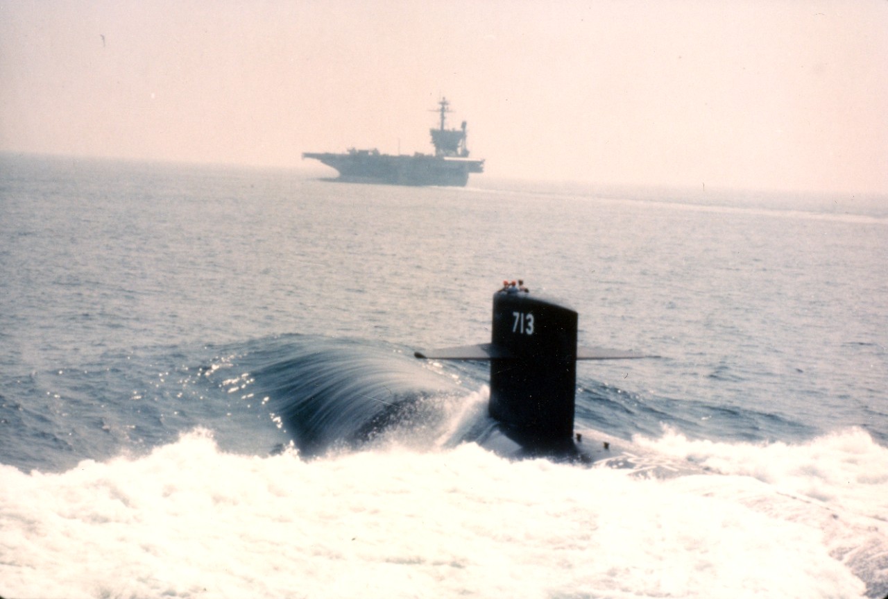 A port quarter view of the nuclear-powered attack submarine USS Houston (SSN-713), foreground, and the aircraft carrier USS John F. Kenney (CV-67), background, departing Hampton Roads for a patrol. August 17, 1982. 