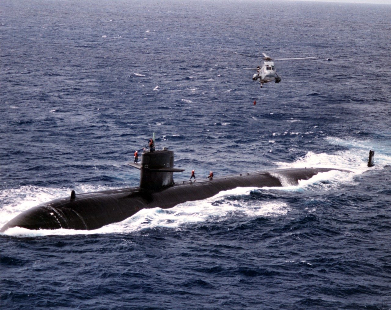 USS Hyman G. Rickover (SSN-709) underway at sea, with an SH-3H Sea King helicopter hovering overhead